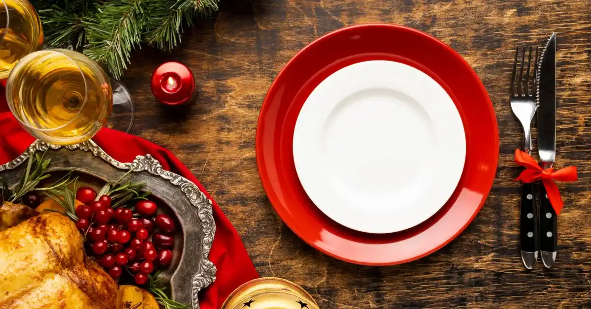 Festive table setting with an empty white plate on a red charger, roasted chicken with cranberries, wine glasses, cutlery tied with a red ribbon, and holiday decorations on a wooden table.