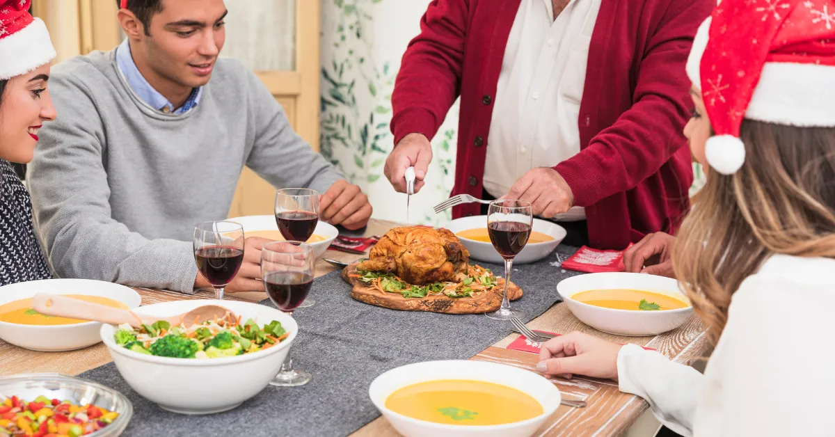 Family enjoying a summer dinner together, carving roasted chicken at a festive table with soup, salad, and glasses of red wine.