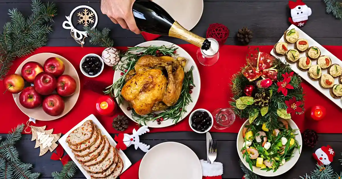 Festive Christmas dinner table with roasted chicken, apples, salad, appetizers, bread slices, and champagne being poured, decorated with red candles, pine branches, and holiday ornaments.