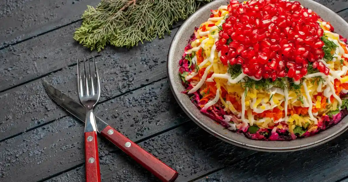Layered vegetable salad topped with pomegranate seeds and fresh herbs, served on a plate with fork and knife on a dark wooden table.