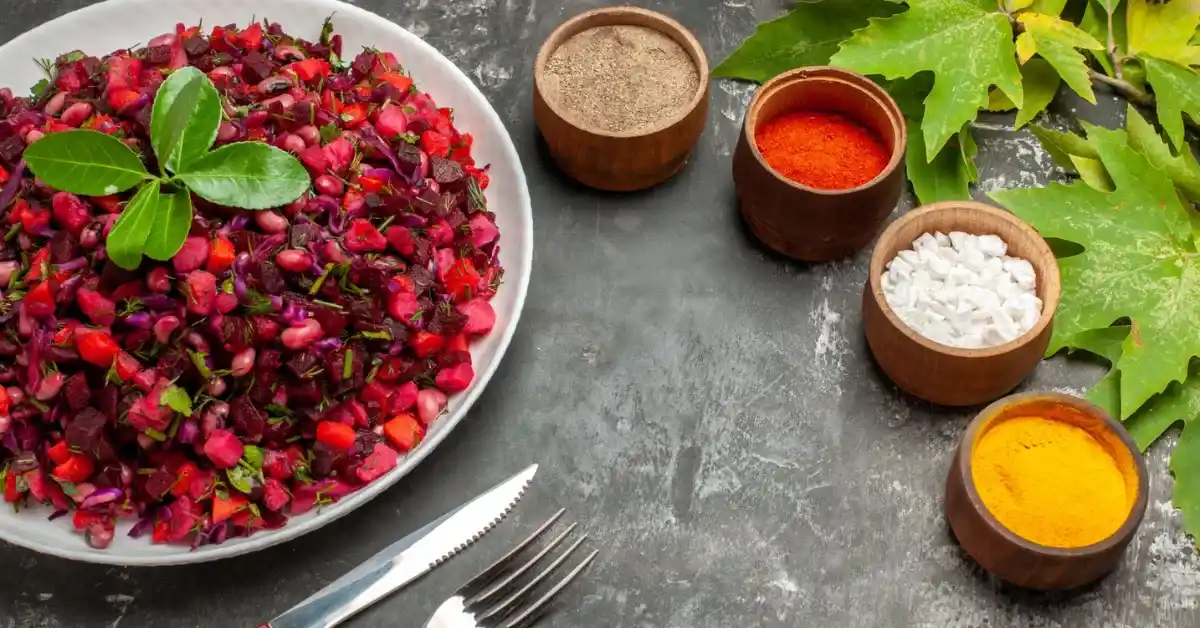 Bowl of fresh beetroot salad with beans and vegetables, garnished with green herbs, served on a grey table with spices and cutlery.