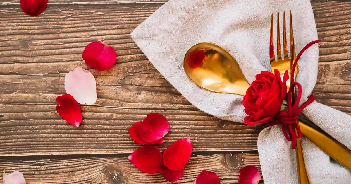 Romantic table setting with red rose petals, golden spoon and fork on a napkin tied with a red ribbon and rose on a wooden background.