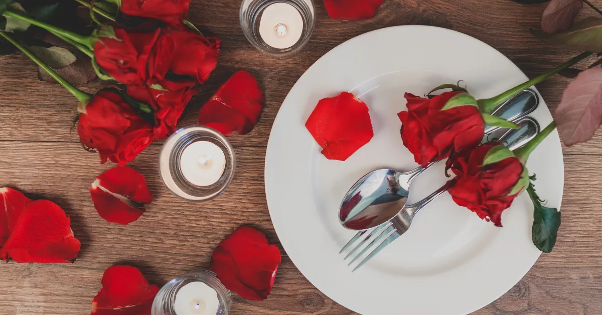 Romantic dinner table setup with red roses, rose petals, candles, and a white plate with spoon and fork on a wooden background.
