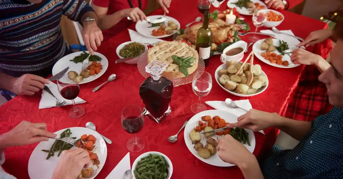 Family enjoying a festive dinner together at a red tablecloth dining table with roasted turkey, vegetables, wine, and pie.