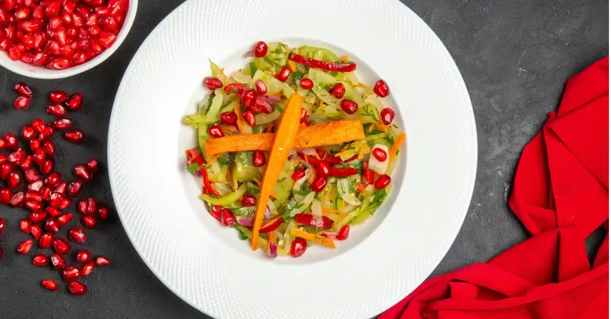 Alt Text: Fresh vegetable salad topped with pomegranate seeds and carrot strips served in a white plate, with scattered pomegranate arils and a red cloth on a dark background.