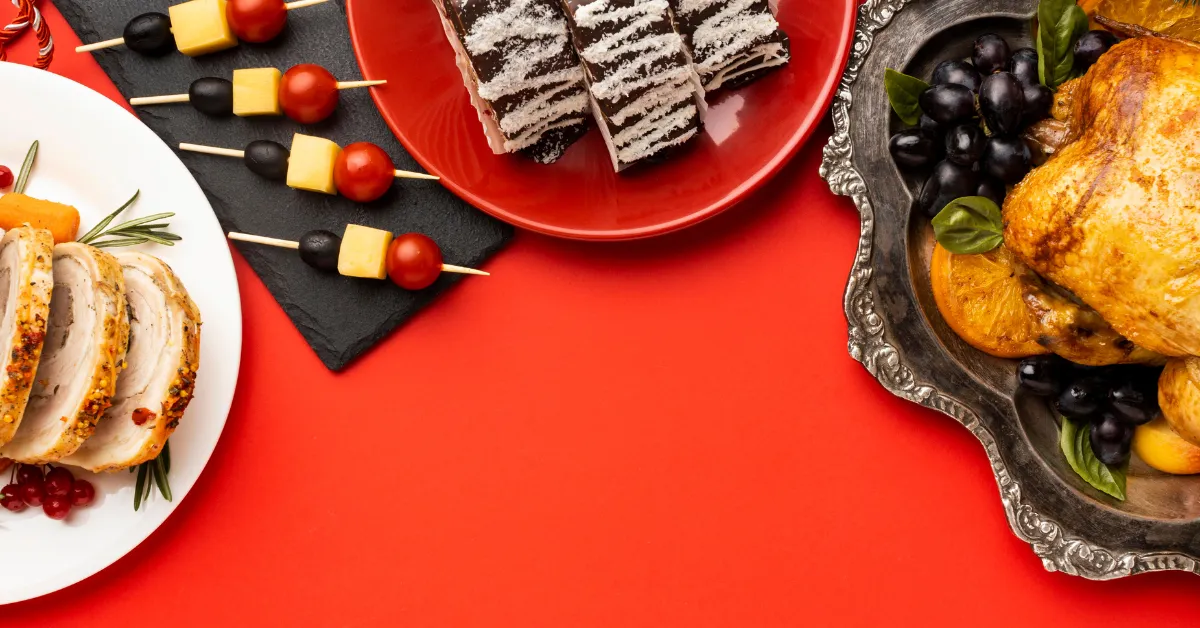 Festive holiday food spread with roasted chicken, sliced turkey, olives, cheese skewers, and layered cake on a red background.