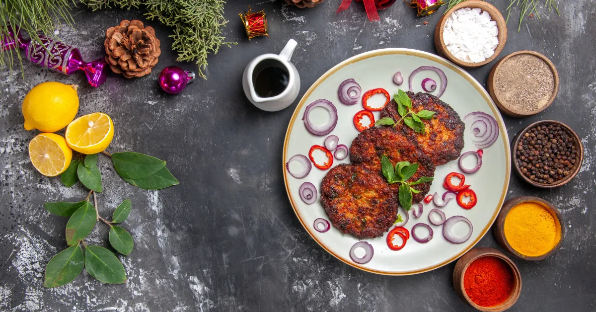 Top view of crispy fried patties served on a plate with sliced onions, red chili, fresh herbs, and surrounded by spices, lemon, and seasoning ingredients on a rustic table.