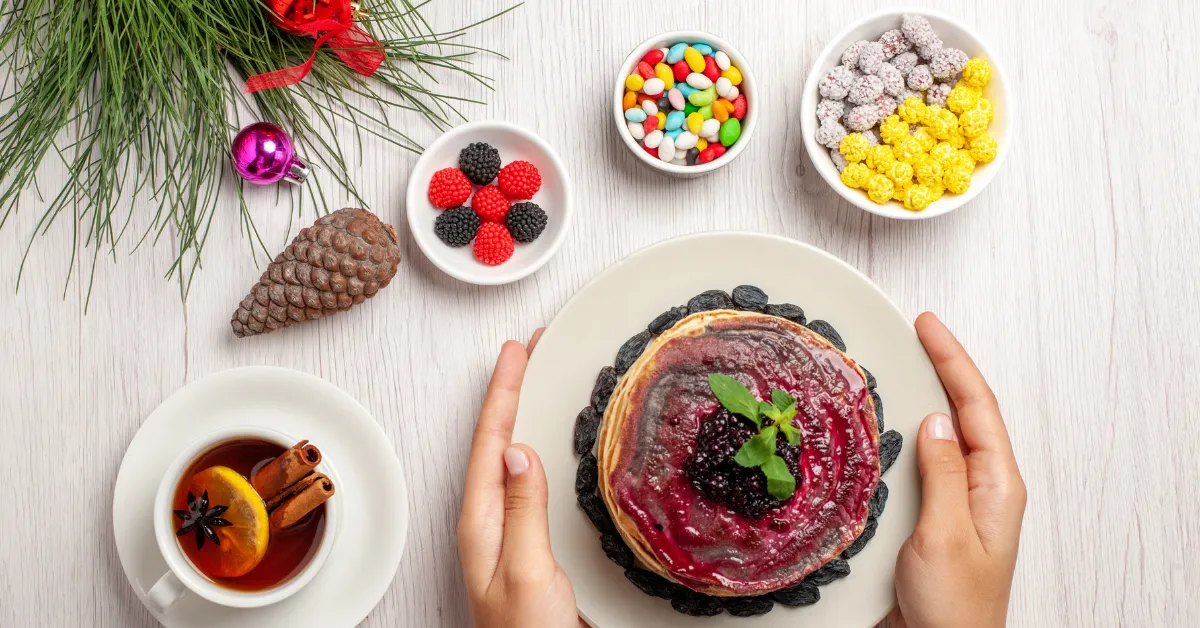 Hands holding a plate of pancakes topped with berry sauce and mint, surrounded by colorful candies, popcorn, tea with cinnamon, and holiday decorations on a wooden table.