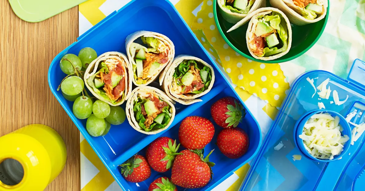 A colorful lunchbox with tortilla wrap pinwheels filled with vegetables, served alongside green grapes and fresh strawberries, with a side of shredded cheese.