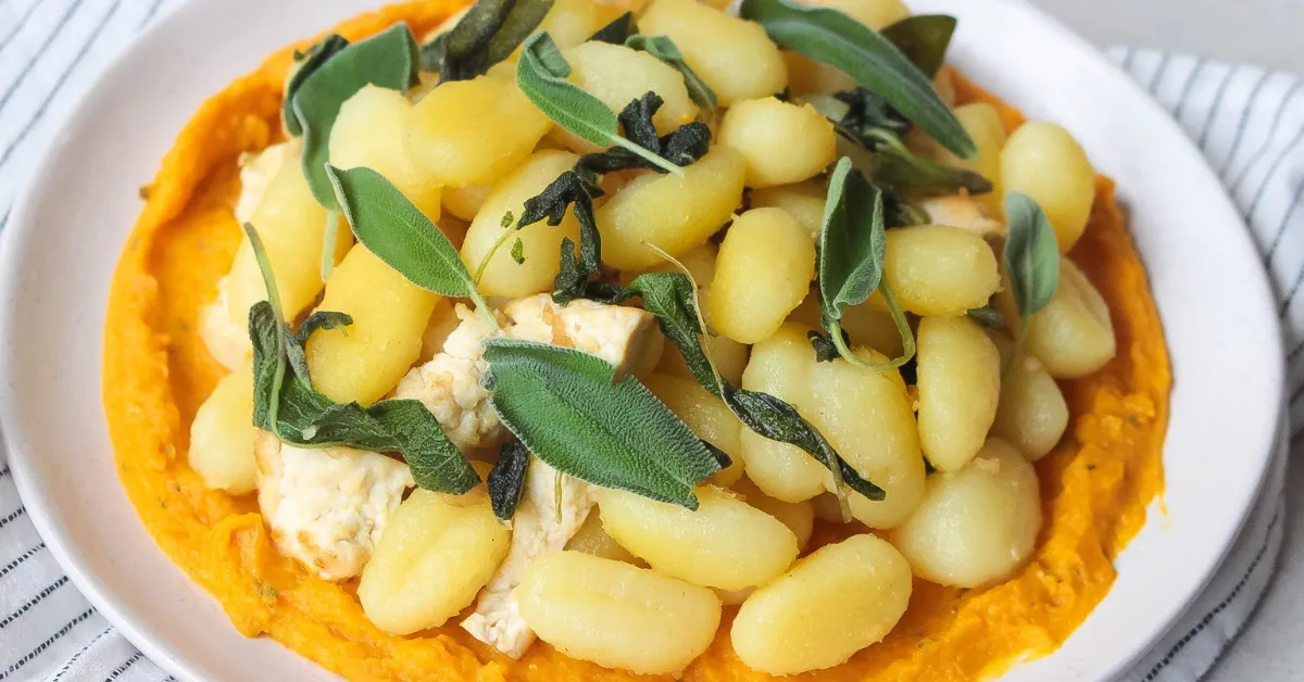A plate of potato gnocchi served over a creamy orange puree, topped with soft cheese and garnished with fresh sage leaves.