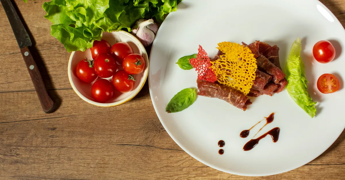 Gourmet plated meat dish with decorative crisp garnish, cherry tomatoes, lettuce, and sauce on a white plate with fresh ingredients on a wooden table.