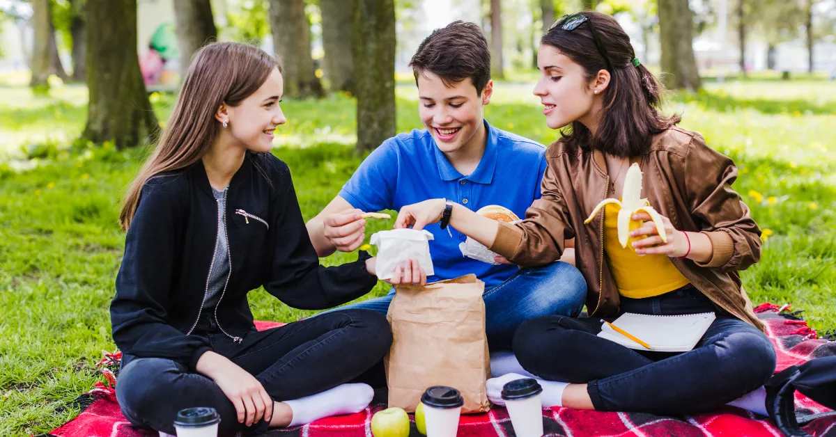 Three teenagers sitting on a picnic blanket in a park, sharing snacks and fruit while enjoying an outdoor lunch together.