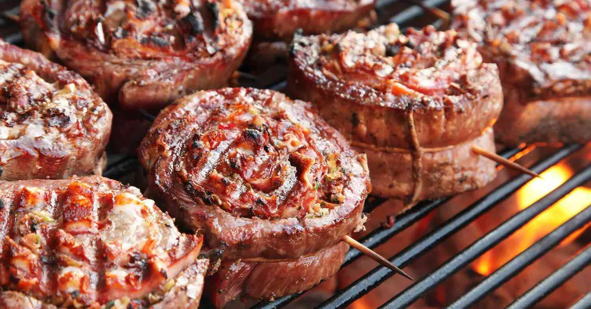 Grilled beef pinwheel steaks on skewers cooking over an open flame with visible char marks and melted seasoning.